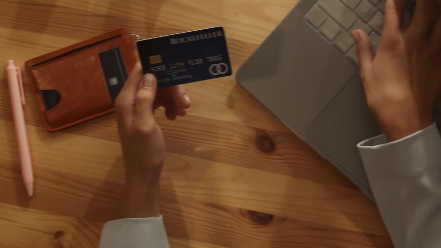 The video shows an overhead view of a person preparing to make an online payment. They place a credit card labeled rockefeller next to a wallet and a sticky note reading pay contractor then begin typing on a laptop