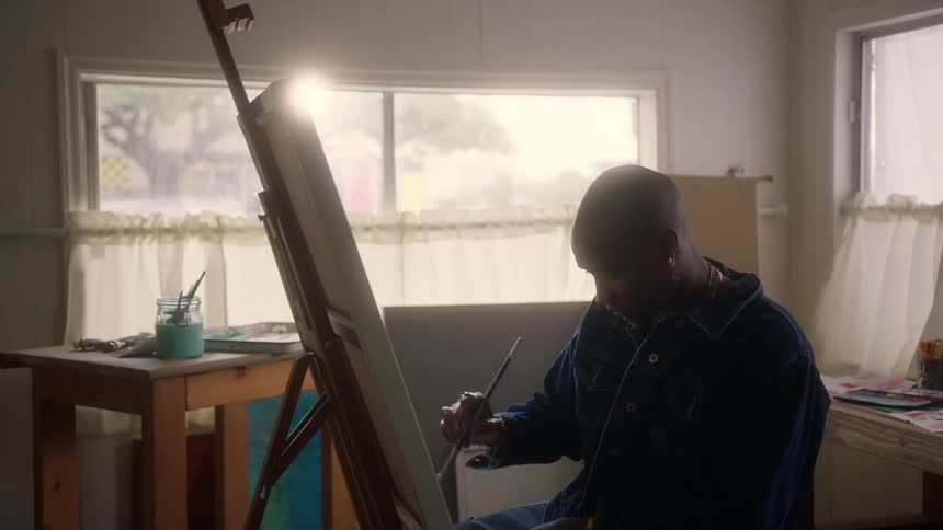 An artist paints on a canvas in a sunlit studio with a large window