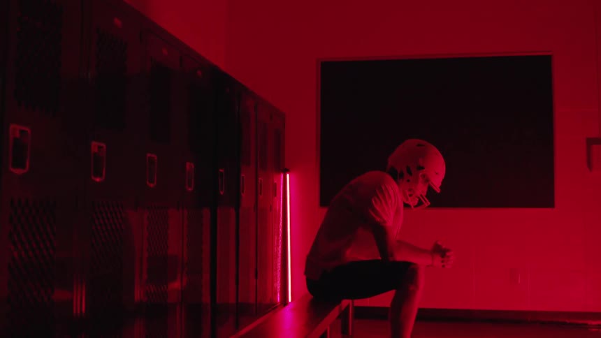 American football player sits in the locker room under intense red lighting