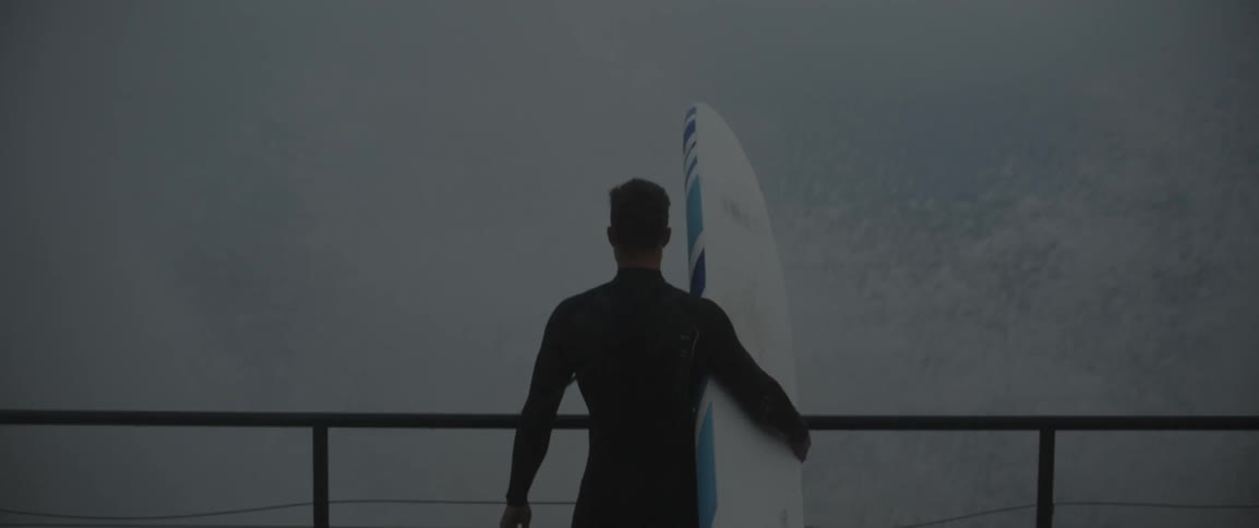 Man holding surfboard standing on walkway watching crashing waves