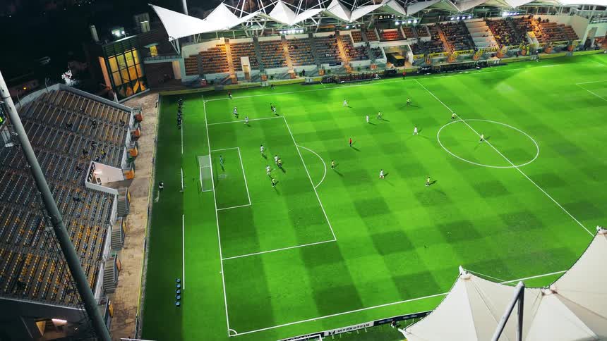 Bright lights illuminate a football field during a match with players running across the pitch as fans watch from the stands