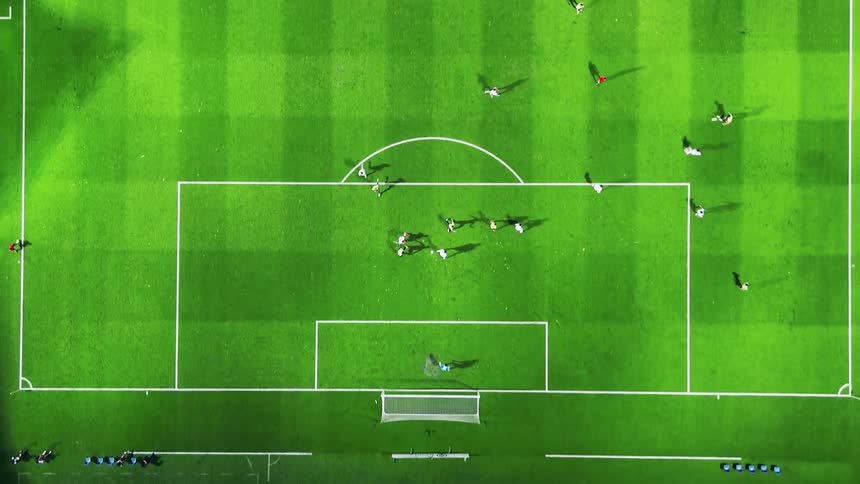 A soccer match in progress on a green field clearly marked with white lines featuring multiple players moving the ball and casting long shadows as the camera slowly tracks from the center circle towards the goal box