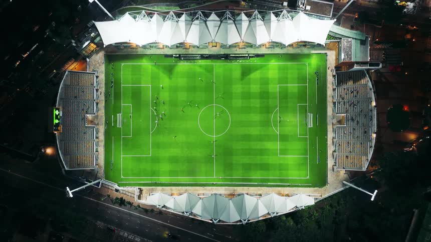 An overhead highangle drone shot captures an illuminated football stadium at night centered on the bright green pitch where small figures of players are active surrounded by dark seating and a white angular roof structure