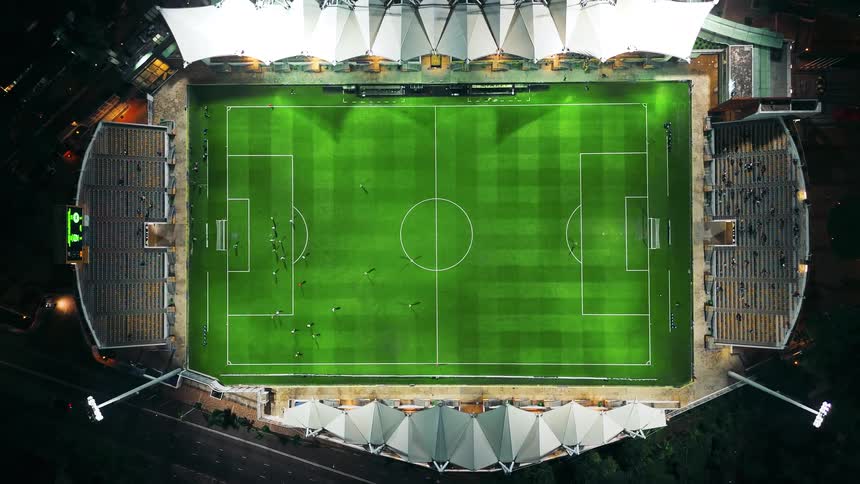 An aerial shot begins focused on the center of a brightly illuminated green soccer field with players moving on it and then the camera slowly zooms out to reveal the entire stadium structure at night including the surrounding areas