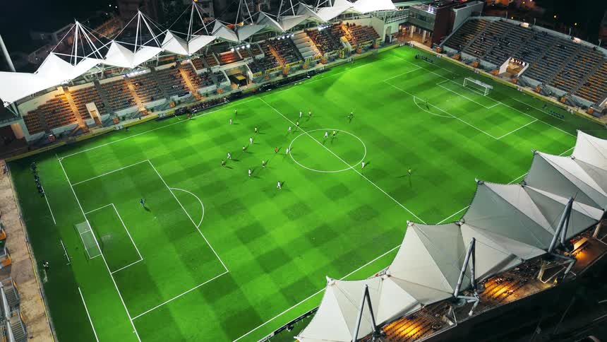 An overhead view captures a soccer match being played at night on a brightly lit green striped stadium field surrounded by orange seats and a complex roof structure
