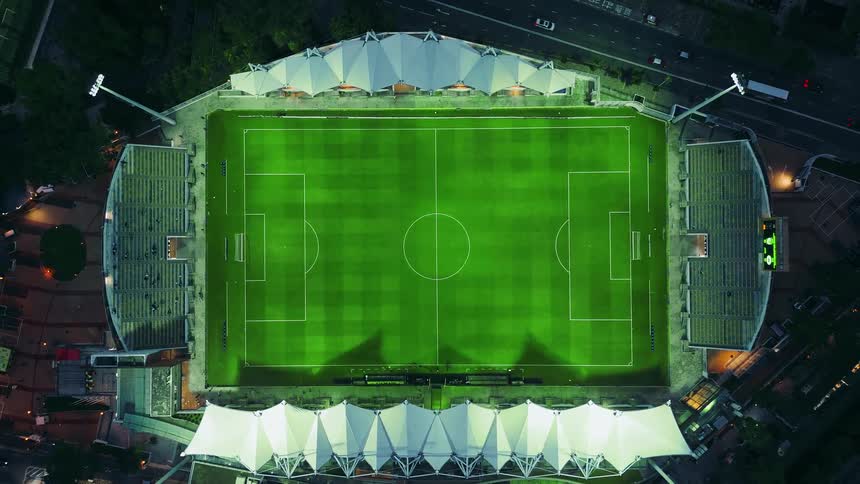 A few people sit in the stands surrounding one of the soccer fields located near a highway at night