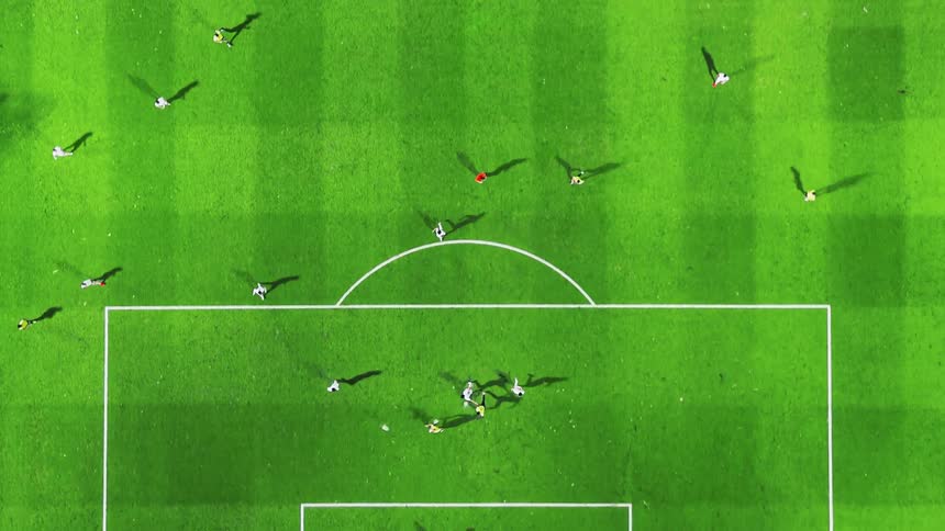 An overhead view captures a soccer match being played on a bright green pitch with players running and casting long shadows in the sunlight
