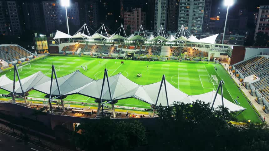 An aerial wide shot shows a brightly floodlit stadium with a distinctive white tensile roof structure and a green sports field where a game is underway surrounded by tall urban buildings at night while traffic moves on the adjacent road