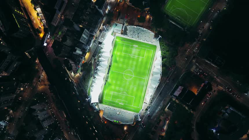 A stadium with a brightly illuminated green soccer field surrounded by dark urban buildings at night slowly rotates and zooms out to show the entire whiteroofed structure and the surrounding city block