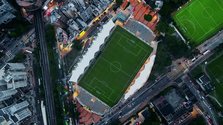 This is a wide aerial shot capturing a soccer stadium surrounded by a dense urban environment at dusk or nightthe stadium field is brightly lit and empty and the surrounding streets show moving traffic and brightly lit buildings