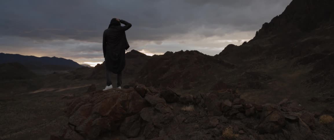 A man stands on a rocky ledge and looks at hills and sky