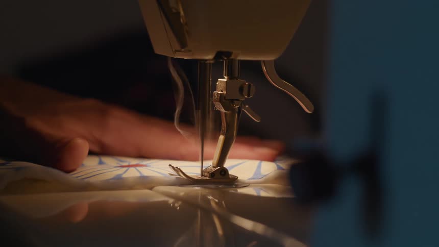 Hand guides patterned fabric under sewing machine needle in operation lit by warm light