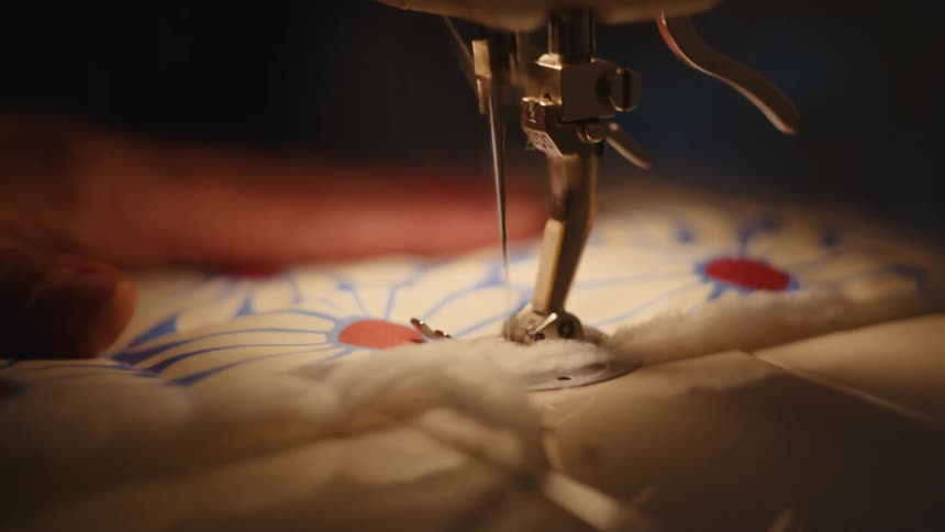 Needle pierces floral fabric with white and red flowers under metal presser foot of sewing machine