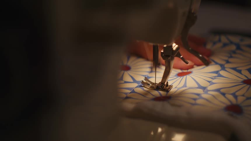 The presser foot and needle of a sewing machine repeatedly moving up and down to stitch a piece of quilted fabric with a blue and yellow daisy pattern while the persons fingers gently guide the material from the right