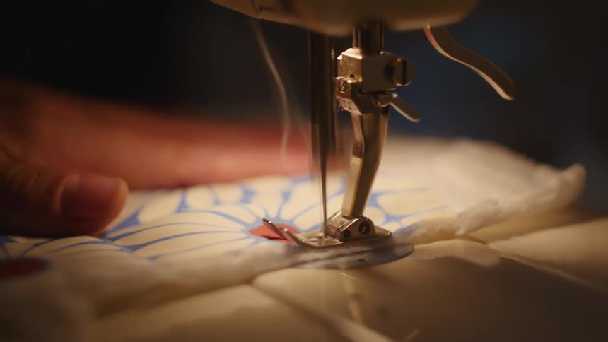 A closeup shot focuses on the metal presser foot and needle of a sewing machine as it rapidly stitches through a thick piece of patterned quilted fabric