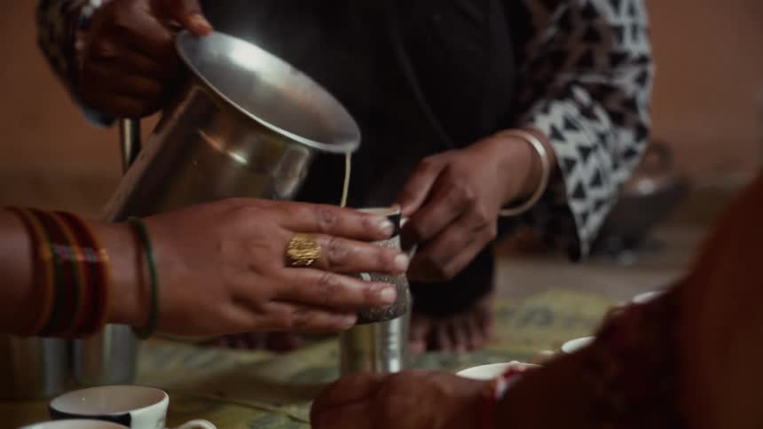 A woman uses a large metal pitcher to pour a lightcolored liquid into a small dark patterned cup being held by another person while several other small cups rest on the table in front