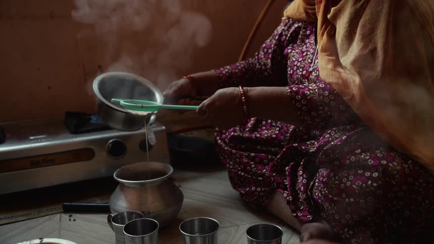 Adult asian woman wearing an orange sari and a pink dress sitting on the floor preparing tea with another person sitting next to her indoors during day time