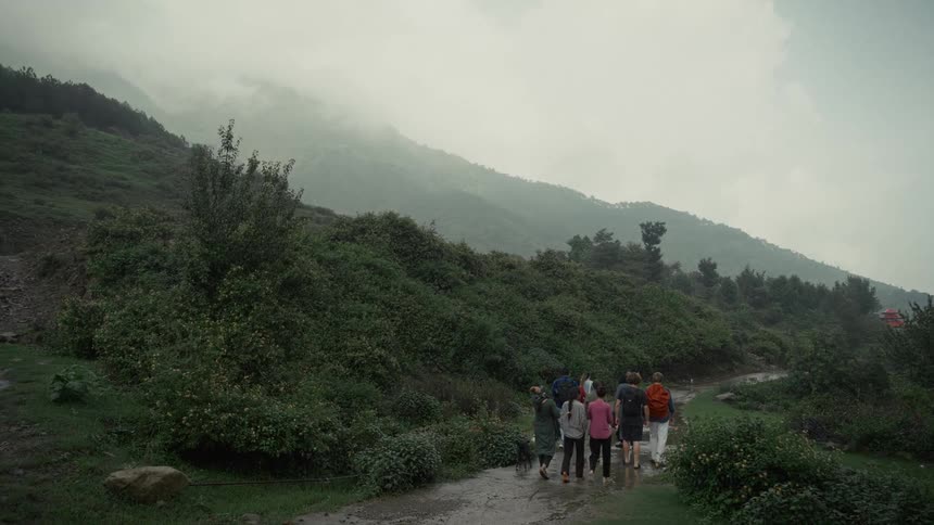 A group of hikers walks along a wet dirt path through foggy green mountains