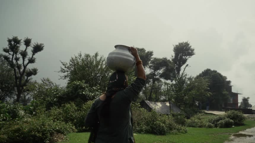 Woman carrying a pot on her head walks along a path in a rural town on an overcast day