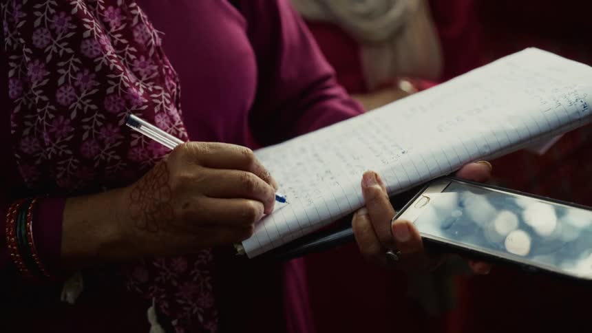 A close view shows a person wearing traditional patterned clothing writing notes on a pad of paper while holding a smartphone below it