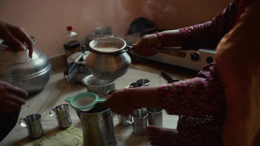 Person straining a hot beverage into a metal container while utensils are spread around