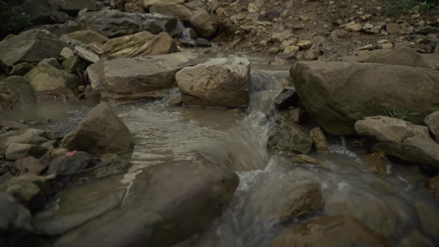 A handheld shot captures a small murky stream flowing steadily over a bed of rough grey rocks