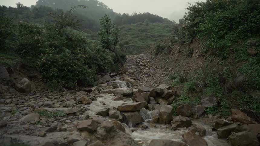 The short video shows a stream of water flowing rapidly over a rocky uneven riverbed in a hilly forested areathe banks are steep and covered in dense green vegetation with the overall scene appearing misty or overcast