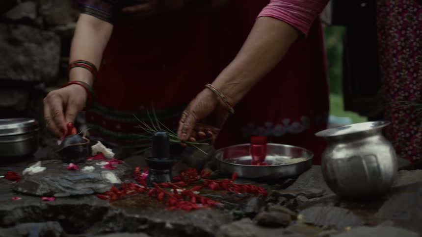 The video shows a closeup of two people performing a religious ritual outdoorsthey are placing offerings including red flowers and other items on a small stone platform that includes a small shiva linga