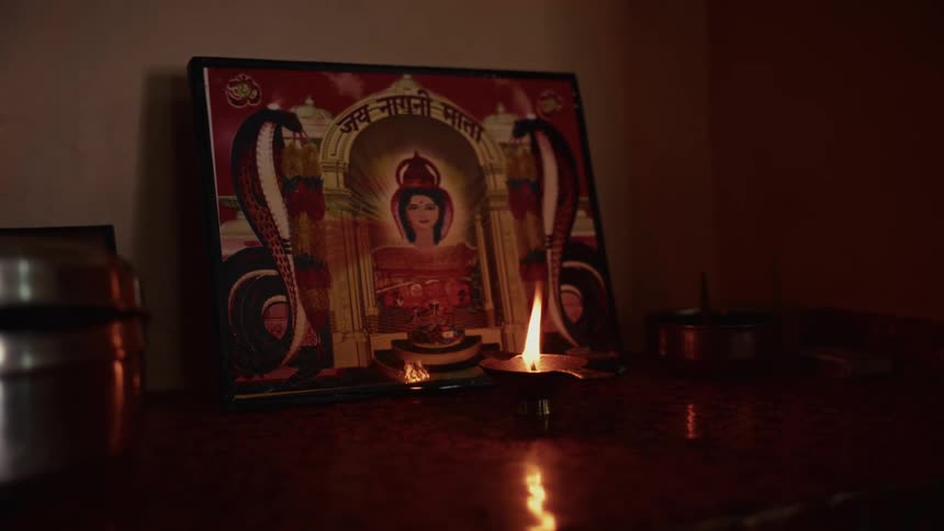 A framed hindu deity print featuring a female goddess and two cobras is displayed next to a lit traditional oil lamp creating a dark devotional atmosphere