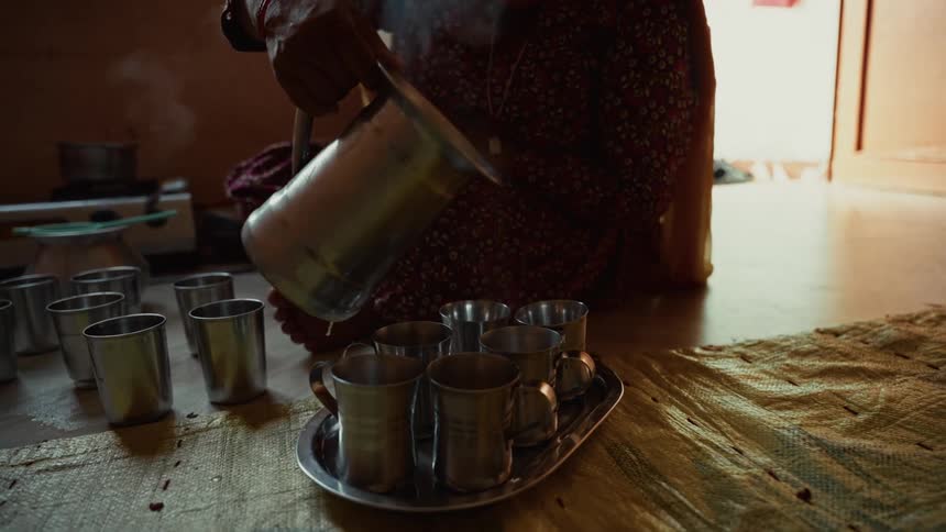 A closeup shot of a persons hands holding a large silver pitcher and pouring a hot steaming dark liquid into several small metal cups arranged on a serving tray while seated on a floor mat indoors