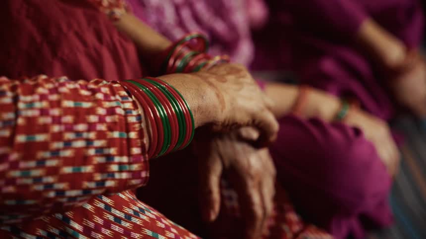 The hands and forearms of a woman wearing numerous red and green bangles and the sleeve of a red patterned garment while the hands rest together near another person dressed in purple clothing