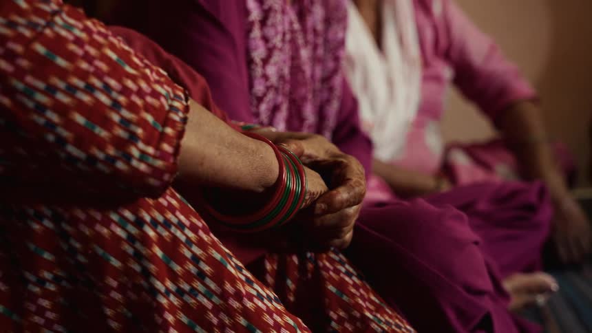 The hands of a woman wearing multiple red and green glass bangles and a red patterned garment with her hands resting on her lap next to another person wearing purple clothing and others blurred in the background