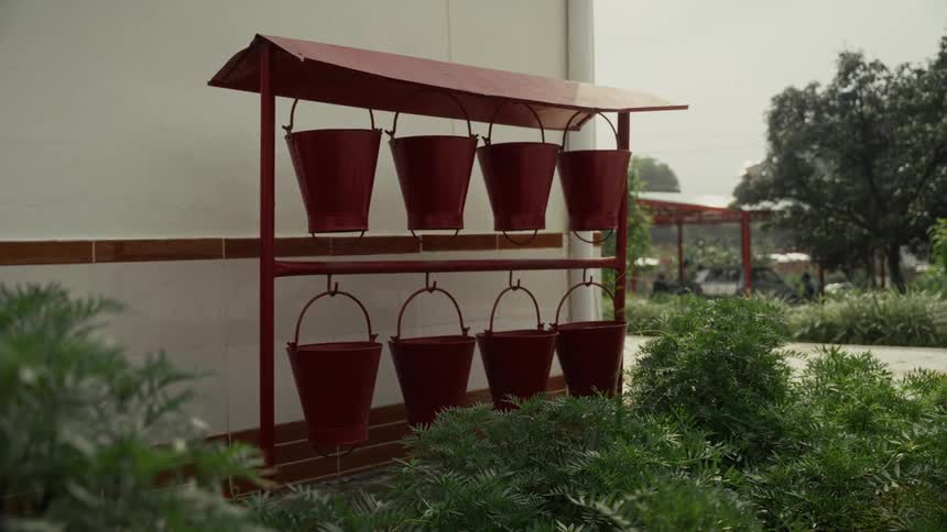 A metal rack holding eight red fire buckets is situated outdoors next to a white wall and green landscaping