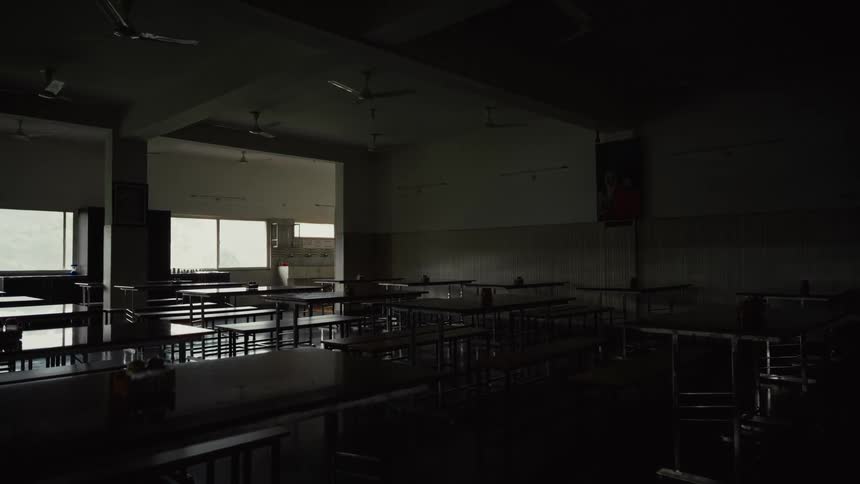 A wide shot of a dark and empty cafeteria or dining hall shows rows of tables and benches with bright daylight coming from windows on the left wall