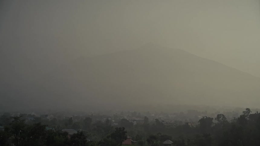 A wide static shot captures a landscape dominated by a large featureless mountain obscured almost entirely by thick gray mist or haze with the low visibility barely revealing the rooftops and trees of a small residential town