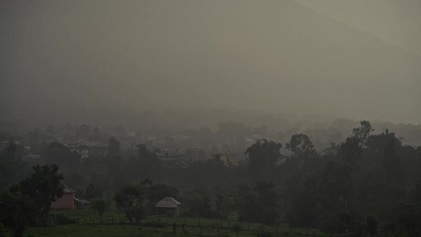 A wide overcast shot of a village nestled in a valley with lush dark green trees and buildings obscured by thick fog or haze