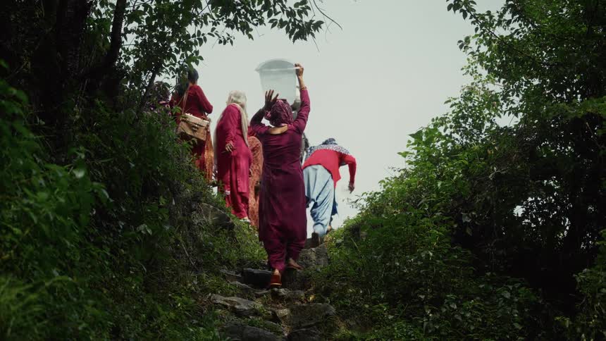 A group of women dressed in colorful traditional clothing like salwar kameez are walking up a steep narrow rocky path the woman in the center is carrying a large transparent plastic bucket of water balanced on her head
