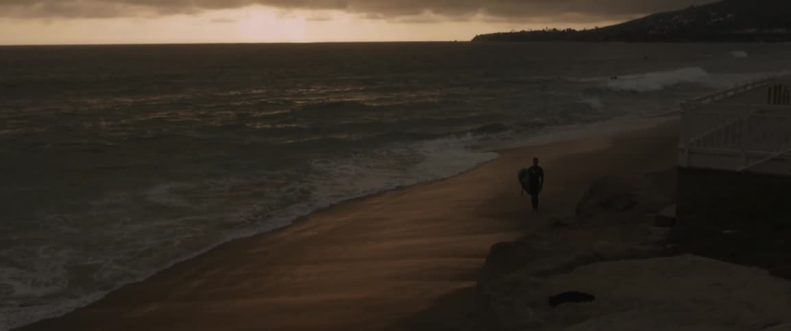 A man walks along the shore at sunset with his board