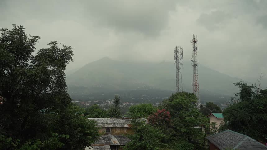 A static wide shot captures a hillside village featuring two tall communication towers set against a backdrop of a massive fogshrouded mountain under an overcast sky
