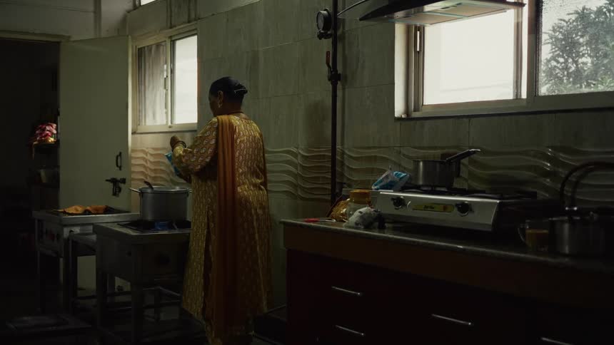The video shows a medium shot of a person seen from the back wearing a traditional yellow and orange patterned dress standing in a dim kitchen they appear to be stirring a pot on a stove in the foregroundthe kitchen has two windows one open to th