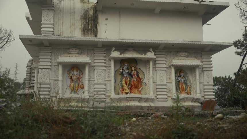A white stone hindu temple with carved niches and colorful statues stands under a gray sky