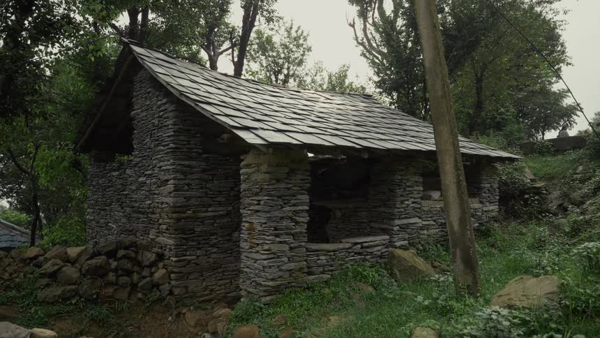 A small rustic building with stacked stone walls and a steep dark slate roof stands amidst thick trees and overgrown green vegetation on a hillside under a pale sky