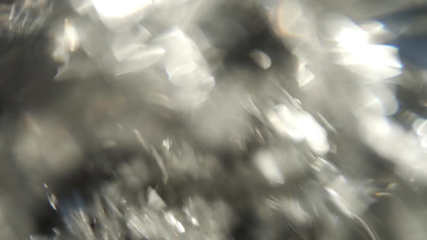 This video shows an abstract dynamic closeup of light reflecting off a highly textured dark and clear surface resembling liquid or ice the light sources create bright shimmering flares and bokeh effects giving the footage a chaotic energetic and