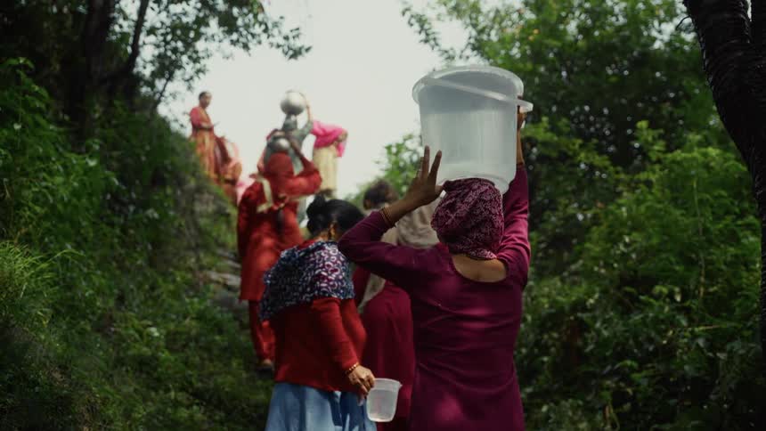 A line of women in traditional clothing and head coverings carry water vessels up a steep winding path through a dense green hillside