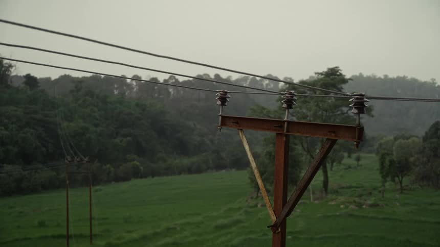 Rusty utility pole with power lines crosses a green rural landscape under a cloudy sky