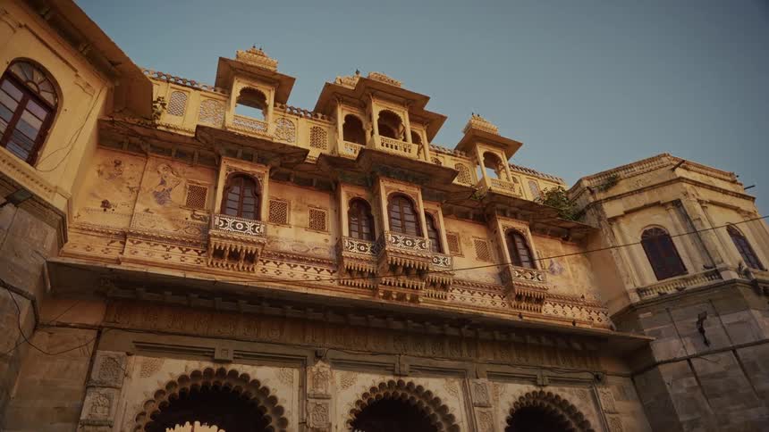 A historic indian palace featuring numerous ornate windows and tiered balconies above large intricately carved arched entryways all viewed from a low angle against a muted light sky