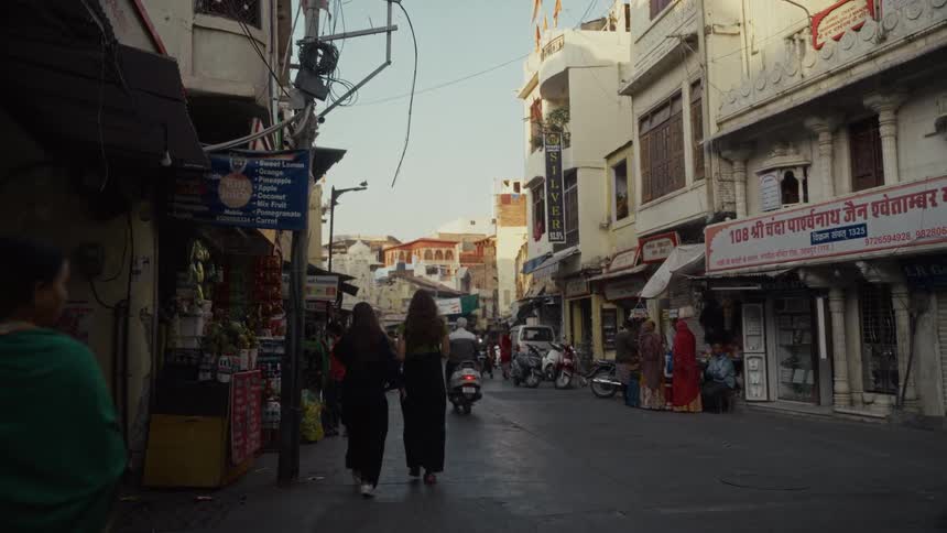 Two women walk along the sidewalk at midday accompanied by another woman as a man rides a scooter past them on the street
