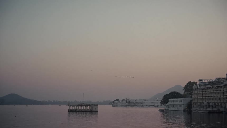 Pavilion on water reflects soft light beside classical buildings and hills in the background
