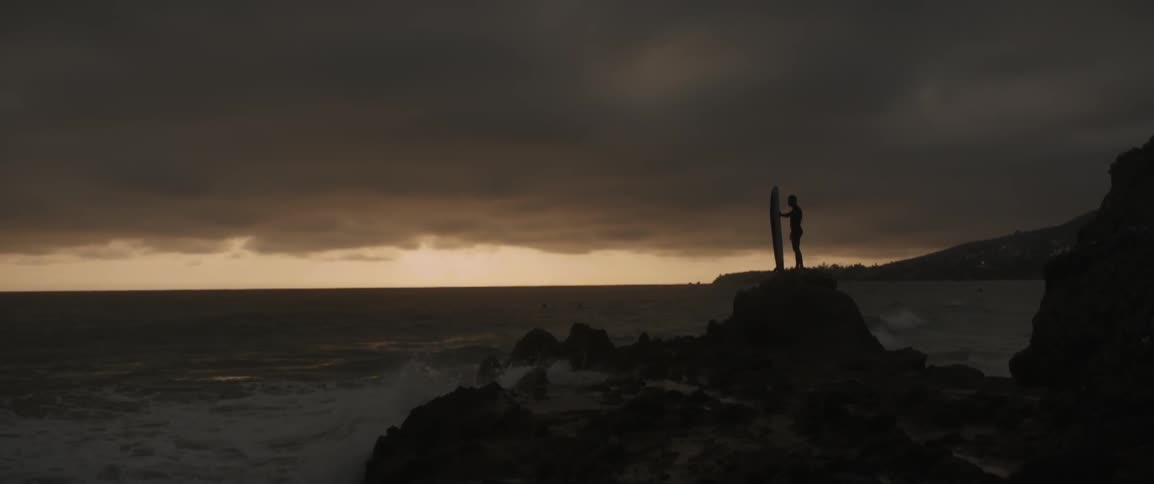 A man is holding a surfboard on some rocks by a rough shore at sunset