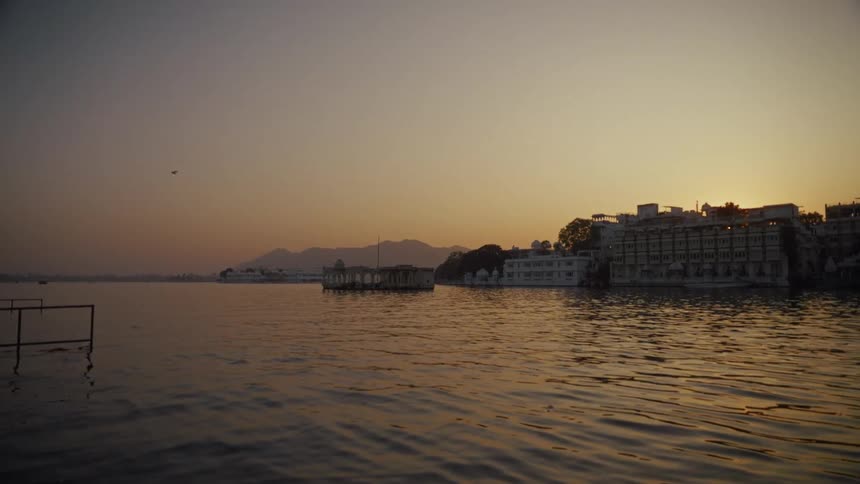 An evening view of a calm lake with traditional white buildings and a small pavilion structure on the distant shore under a glowing orange sky